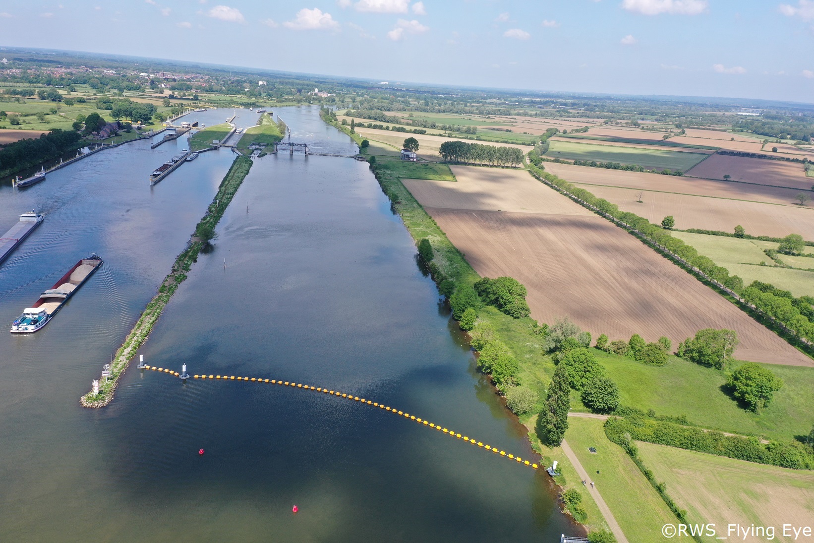 Geul Ossenkamp met een stuwdam onderin en boten die varen in de Maas links. Rechts grenst het water aan een boerenlandschap.